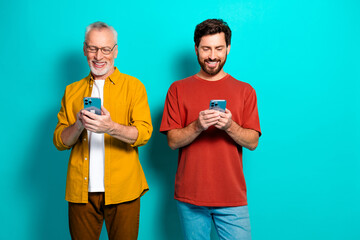 Two men smile and use smartphones together in a casual friends and family lifestyle shot against a teal background