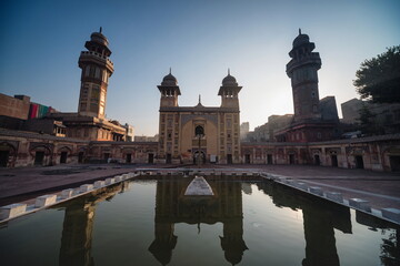 View of the serene, reflective pool mirroring the golden facade of the historic mosque, flanked by towering minarets under a vast sky, Lahore, Punjab, Pakistan.