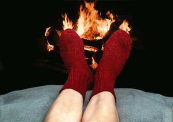 Winter image with relaxed legs in bright red knitted socks near burning fireplace. Warmth, comfort.