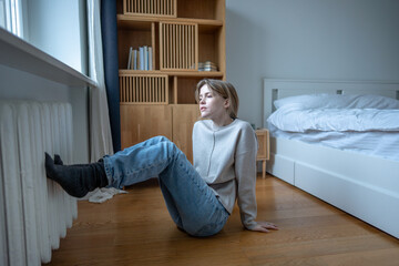 Woman sits on bedroom floor warming feet against heater, struggling with poor home heating during cold season. Frozen female pressing feet to central radiator by window in apartment, to escape cold.