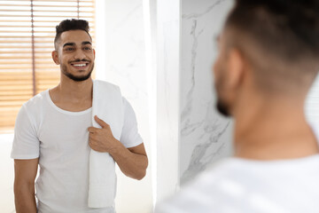 Obraz premium A young man stands in a contemporary bathroom, smiling at his reflection in the mirror. He holds a towel over his shoulder, enjoying a moment of relaxation after showering.