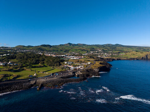 Aerial view of rugged coastline meeting the deep blue sea, with the town nestled amidst green hills under a clear sky, Capelas, Azores, Portugal.