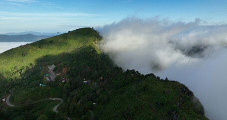 Aerial view of a lush green mountain peak piercing through a sea of white clouds, with a winding road snaking up its side, Tambon Tap Tao, Chang Wat Chiang Rai, Thailand.