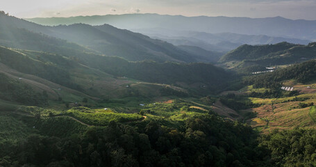 Aerial view of sun-kissed mountains cascade into valleys, where shadows dance amidst lush greenery and scattered settlements, Tambon Tap Tao, Chang Wat Chiang Rai, Thailand.