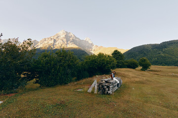 Fototapeta premium Mountain meadow with woodpile and person sitting near trees, landscape view of nature and hiking trail at sunrise, peaceful alpine field and rustic outdoor scenery.