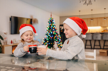Two children in Santa hats happily drinking a hot beverage from mugs by the Christmas tree, holiday tradition