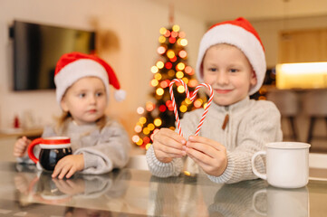 Siblings in Santa hats smiling while sipping from mugs by the Christmas tree, capturing seasonal joy and warmth