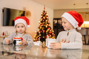 Close-up of two children in Santa hats enjoying a hot drink from mugs near a Christmas tree, winter comfort