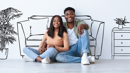 A couple sits on the floor of their living room, smiling and enjoying time together. They are...