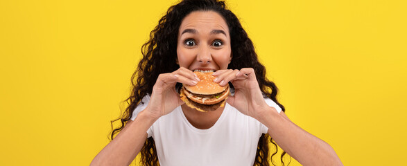 A woman with curly hair smiles widely while holding a big hamburger. The cheerful yellow background enhances her excitement about the delicious food.
