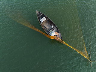 Aerial view of a lone fisherman casts his vibrant yellow net from a small boat on the calm, jade-green waters, Hội An, Quảng Nam, Vietnam.