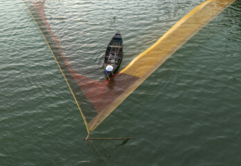Aerial view of a lone fisherman in a small boat navigating the waters, framed by the vibrant yellow and red hues of his fishing net, Hội An, Quảng Nam, Vietnam.