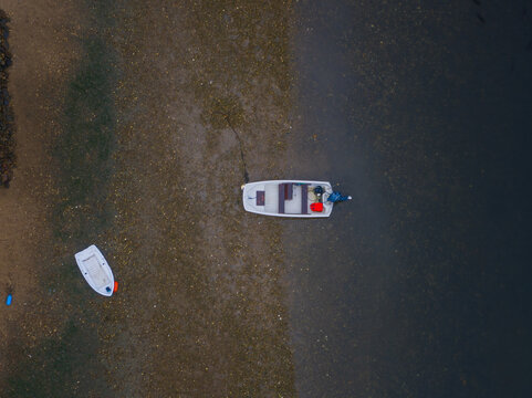 Aerial view of two boats resting on the contrasting textures of wet sand and dark water, a serene tableau of maritime life, Duxbury, Massachusetts, United States.