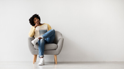 A young black man sits in a cozy chair, resting his feet on the armrest, while contemplating...