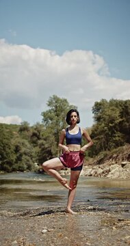 A young woman finds peace and balance practicing tree pose yoga outdoors by a serene river. Her healthy lifestyle is reflected in this mindful exercise, connecting with nature under a sunny sky.