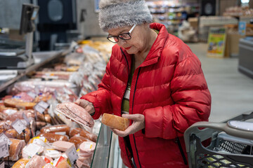 Senior woman reading product label while shopping for deli meat in supermarket refrigerated section.Smart Senior Consumer: Elderly Woman Budget Shopping and Comparing Meat Prices