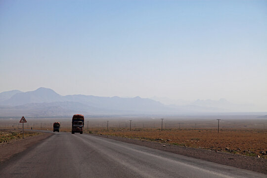 View of two trucks rumble down a sun-baked asphalt road cutting through the vast, arid landscape, with distant mountains fading into the hazy horizon, Quetta, Balochistan, Pakistan.