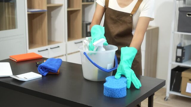An Asian woman cleaner wearing gloves is wiping a computer monitor with a cloth in a modern office. She works carefully and smiles, maintaining cleanliness and hygiene in the workspace