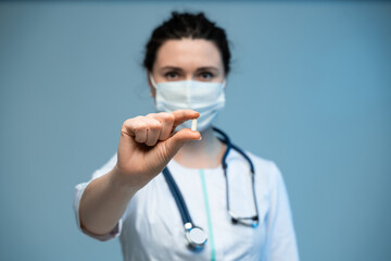 Female doctor in mask holding white capsule toward camera while standing in uniform on blue background