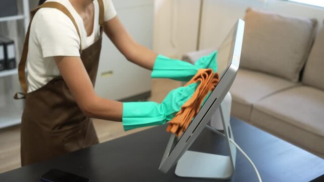 An Asian woman cleaner wearing gloves is wiping a computer monitor with a cloth in a modern office. She works carefully and smiles, maintaining cleanliness and hygiene in the workspace