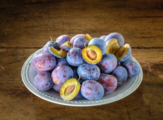 A bountiful collection of ripe, juicy plums, some halved to reveal their golden flesh, rests invitingly on an ornate white plate atop a weathered wooden surface