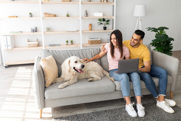 A couple sits together on the couch in their living room, using a laptop. Their happy dog rests nearby, creating a warm and relaxed atmosphere in the home.