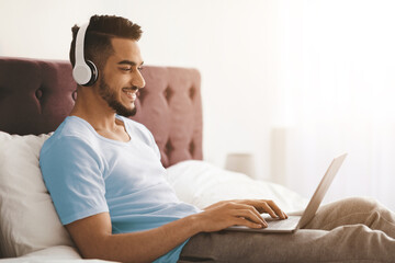 A young man smiles as he listens to music through headphones while using a laptop in his cozy bedroom. Natural light fills the room, creating a warm atmosphere.