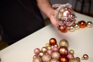 Hands arranging Christmas New Year baubles on white table with tinsel and warm bokeh lights, metallic red gold pink palette, careful styling by home decorator, festive craft mood