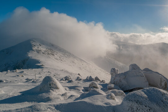 View of snow-covered peaks meeting a blue sky with white clouds, icy rocks scattered across the foreground, Chopok, Banska Bystrica Region, Slovakia.