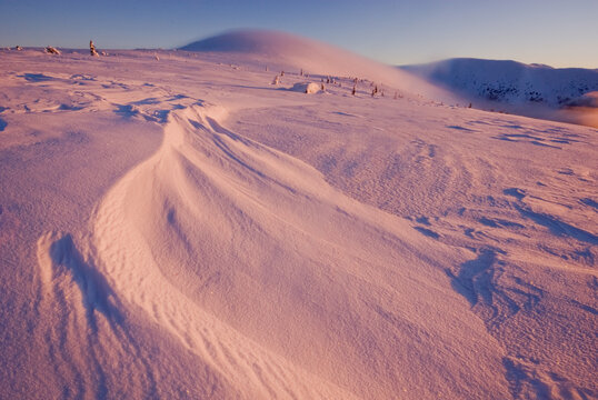 View of the snow-covered landscape, sculpted by wind into smooth curves, glows with the warm hues of sunset, contrasting with the crisp blue sky, Low Tatras, Banska Bystrica Region, Slovakia.