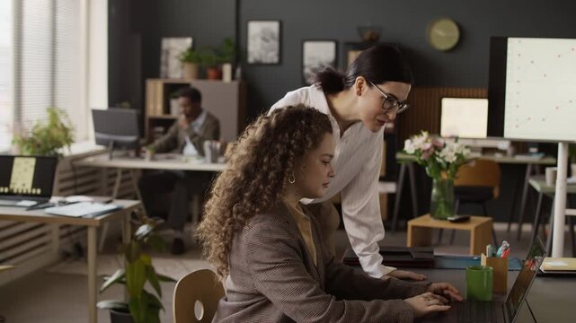 Slow motion of brunette mature woman coming to her curly haired subordinate who working on laptop at desk, they turning heads to camera and smiling