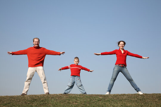 A family of three is standing on a grassy hill, with the man and boy on one side and the woman on the other. They are all wearing red shirts and are stretching their arms out to the side