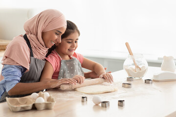 A mother and her daughter are joyfully working together in a bright kitchen. They are rolling out dough, surrounded by baking tools and ingredients, sharing a special moment.