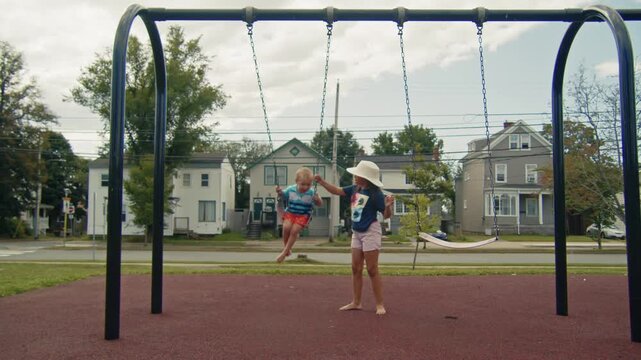 Older Sister Pushing Toddler Brother On Swing In Canadian Suburban Playground. Cute siblings laughing and playing on swings in Dartmouth, Nova Scotia. A sweet family moment captured on a bright summer