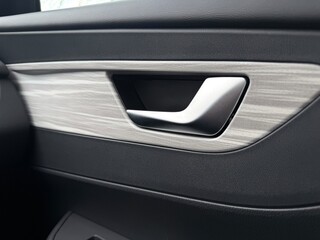 A close-up of a modern car's interior door panel featuring a metallic silver handle recessed into a textured, light gray wood-grain trim. The surrounding material is matte black plastic, highlighting․