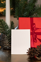 Blank paper card mockup, red gift box, pine cones and branches on table, Christmas holiday postcard or invitation design template with empty copy space, soft focus