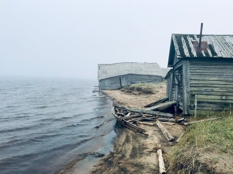 Foggy sandy riverbank with remains of old wooden fishing boats and weathered unpainted boathouses, blackened by time, on the Varzuga River in the Russian North.