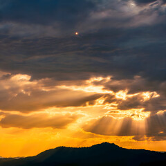 Dramatic sunset sky with sun rays, golden hour mountain silhouette, heavenly light beams breaking through dark clouds, moody orange and yellow atmosphere, nature landscape background, storm weather.