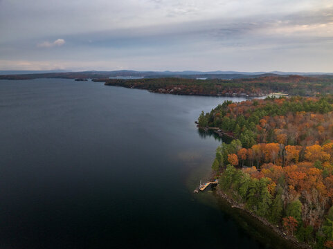 Aerial view of the tranquil lake mirroring the overcast sky, fringed by a vibrant tapestry of autumn foliage, Alton, New Hampshire, United States.