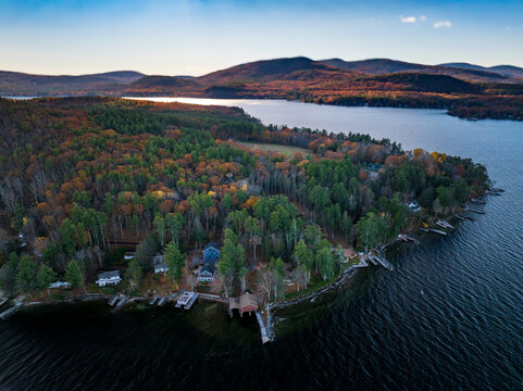 Aerial view of autumnal forests meeting the dark, rippled waters of a lake under a soft sky in Alton, New Hampshire, United States.
