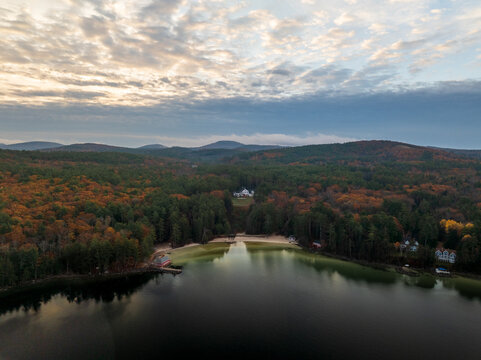 Aerial view of dark waters meeting the autumnal forests, with houses nestled among the trees under a vast sky, Alton, New Hampshire, United States.