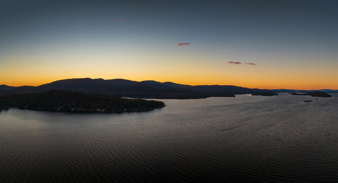 Aerial view of a tranquil lake reflecting the warm hues of the sunset, with distant mountains silhouetted against the fading light, Alton, New Hampshire, United States.