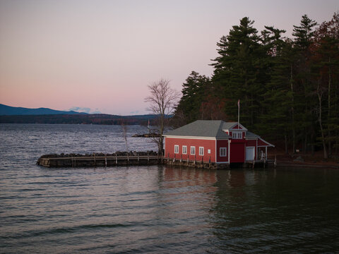 Aerial view of a red boathouse with white framed windows nestled against the dark evergreen trees, extending into the tranquil lake under a soft pastel sky, Alton, New Hampshire, United States.