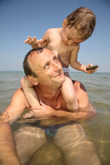 A man senior and a child are playing in the water. The man is holding the child on his back