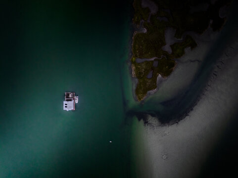 Aerial view of a building sits serenely amidst the turquoise water, while the coastline's sand merges with dark vegetation, creating a stark contrast, Gloucester, Massachusetts, United States.