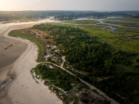 Aerial view of a tranquil landscape where sandy beaches meet lush green forests and winding marshlands under a soft, diffused light, Gloucester, Massachusetts, United States.