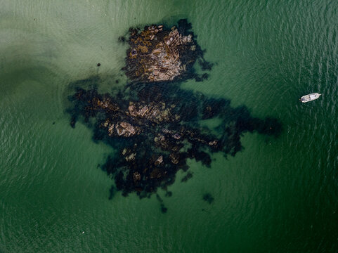 Aerial view of dark rocks contrasting against the shallow, teal waters, near a small boat, Gloucester, Massachusetts, United States.