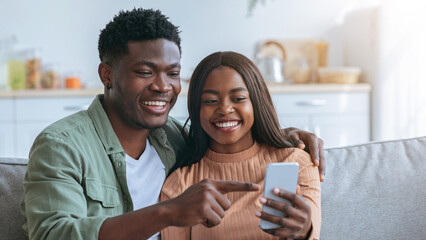 A joyful couple sits closely on a couch, sharing a light moment as they look at a smartphone. The...