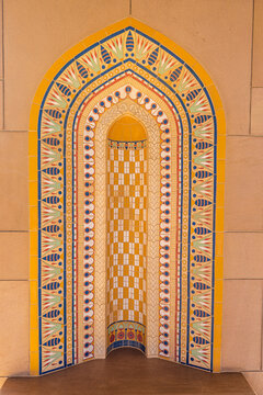 View of an ornately decorated niche, featuring intricate mosaic tilework in shades of yellow, blue, and white, set against a beige wall, Muscat, Muscat Governorate, Oman.