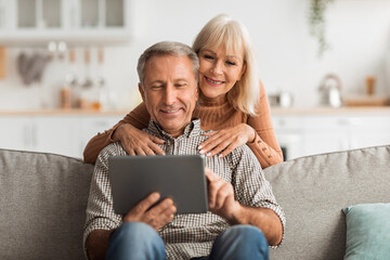 A senior couple sits on a sofa, happily engaging with a tablet in a bright, inviting living room. They share laughter and joy in their warm home environment.